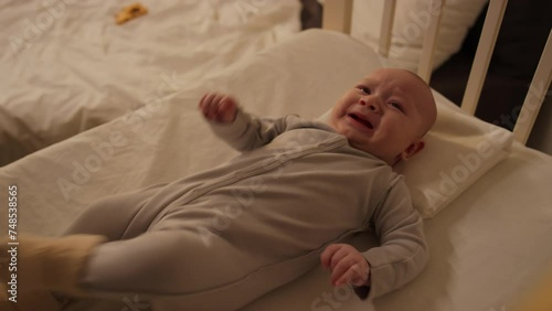 Medium shot of unrecognizable mother or nanny putting cranky baby boy in bodysuit to sleep in crib, and child crying and kicking