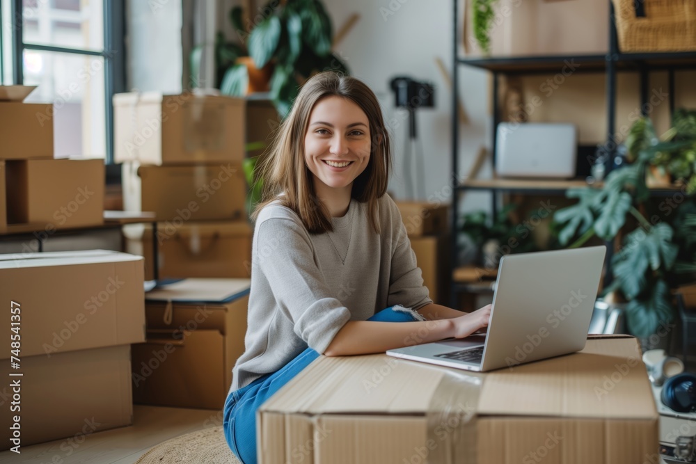Young female entrepreneur smiling at laptop in a home office with moving boxes Young female entrepreneur smiling at laptop in a home office with moving boxes