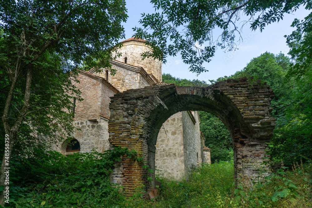 An old stone church with a tiled roof and a dome in the forest. Brick arch of an old wall. There are trees and bushes around. Day, bright blue sky.