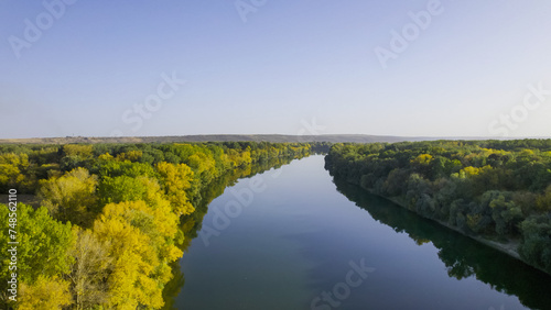 Hovering higher, the drone captures the river bordered by lush yellow-green trees, offering a sweeping vista of nature's seasonal canvas from an elevated perspective