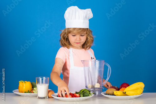 Child chef cook make smoothie prepares food in isolated blue studio background. Kids cooking. Teen boy with apron and chef hat preparing a healthy vegetables meal in the kitchen.
