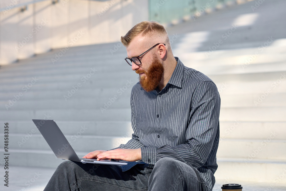 Handsome red-haired freelancer man with a beard sits on the street and works remotely using a laptop