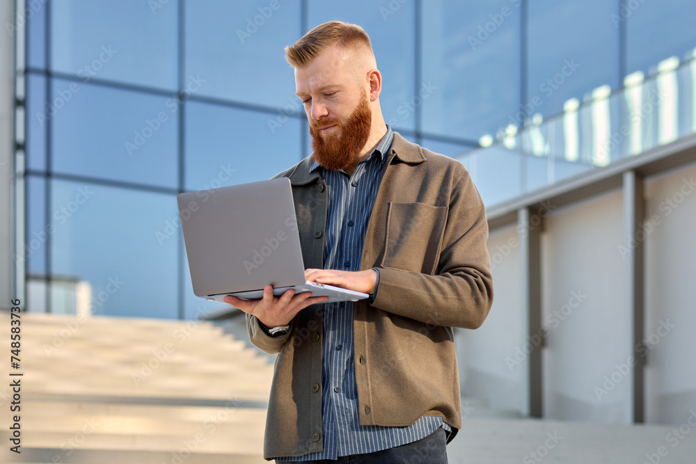 A man with a red beard dressed in a shirt and jacket uses a laptop while standing on the street A man with a red beard dressed in a shirt and jacket uses a laptop while standing on the street
