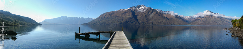Panoramic view of wooden pier jutting into placid and crystal clear Lake Wakatipu towards Cecil Peak in the Southern Alps on New Zealand's South Island