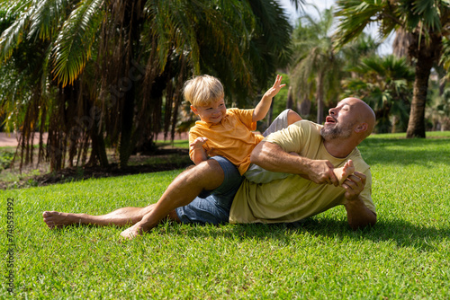 family and love concept - young father tickling his little son in park. dad tickles his son's legs on the grass. dad with little son having fun on the green lawn