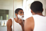 Black man, mirror and shaving with razor in bathroom for grooming, skincare or morning routine. Reflection, beard or person with cream on face for cleaning, health or hair removal for hygiene in home