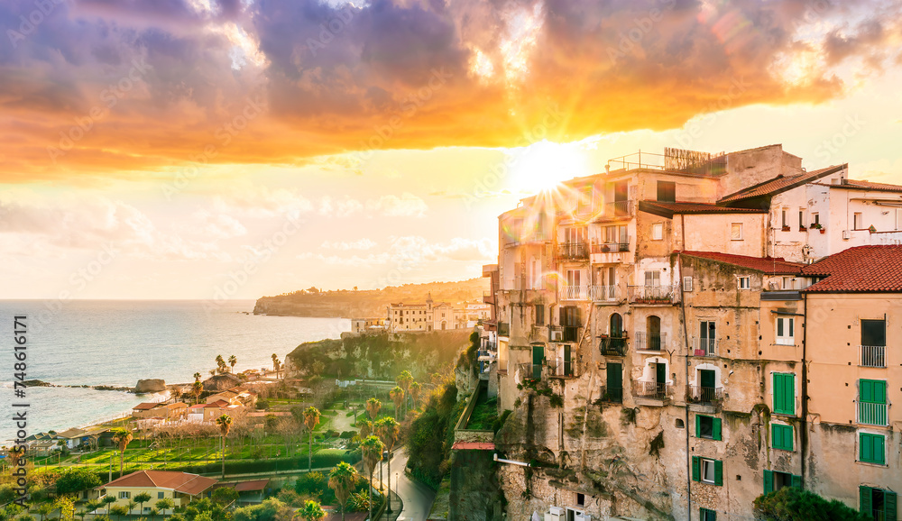 beautiful sunset landscape above sea coastline in Tropea, Italy. Antique buildings on a high rock cliff with green palm trees and scenic cloudy sunset sky on background