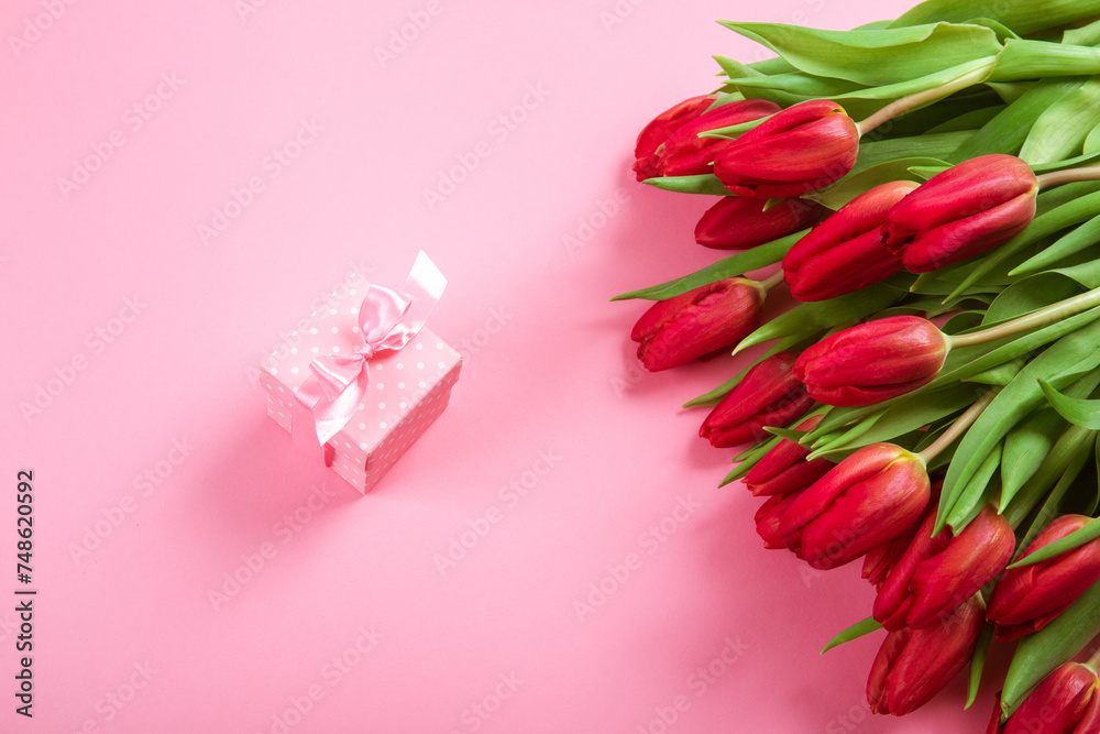 Small pink gift box with a ribbon in white polka color dots with big red tulip bouquet on light pink background. Jewelry gift card certificate. Copy space. Top view. Woman day holiday mockup design Small pink gift box with a ribbon in white polka color dots with big red tulip bouquet on light pink background. Jewelry gift card certificate. Copy space. Top view. Woman day holiday mockup design