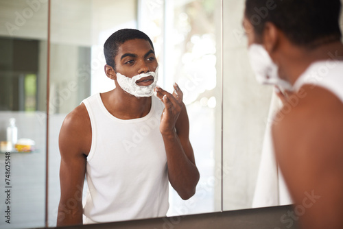 Black man, mirror and shaving cream on face in bathroom for grooming, skincare or morning routine. Reflection, beard and person apply foam for cleaning, health and hair removal for hygiene in home photo