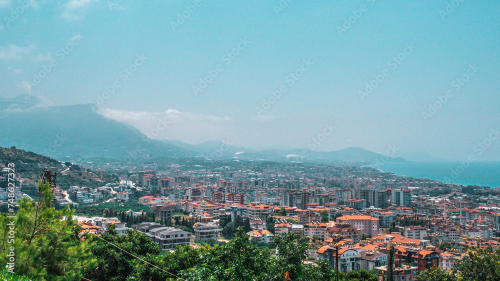 The photo captures the cityscape of Alanya, with the majestic backdrop of a towering mountain, harmonizing urban life with the grandeur of nature's presence, offering a picturesque view of the city's 