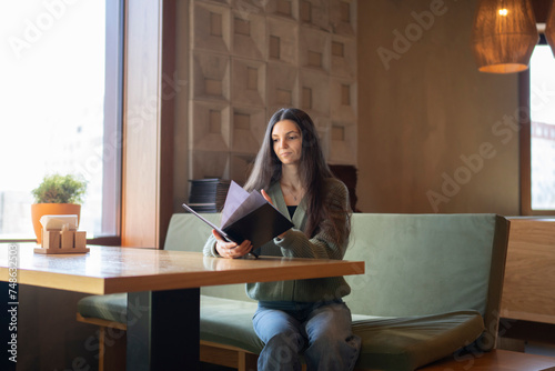 Young Woman Seated on a Green Sofa Choosing a Meal From a Menu in cafe