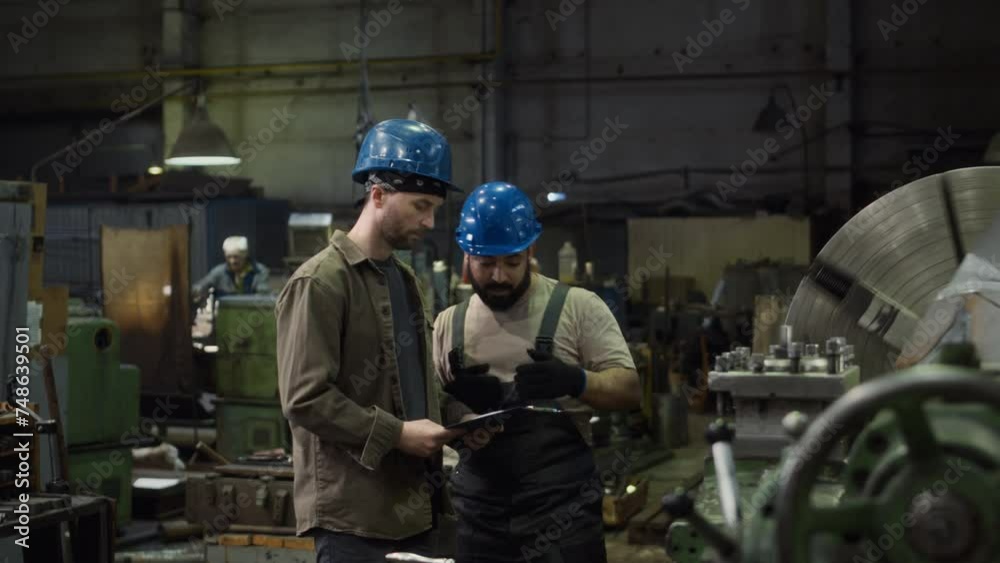 Medium shot of two ethnically diverse male turners in blue hard hats walking along production plant and having discussion while checking work of turning machine using checklist on clipboard Medium shot of two ethnically diverse male turners in blue hard hats walking along production plant and having discussion while checking work of turning machine using checklist on clipboard