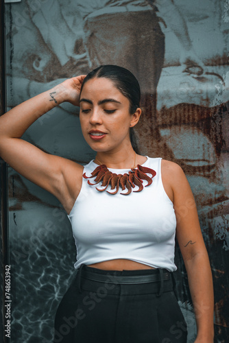 Young woman with long dark hair wears white tank top decorated with feather patterns. Warm smile and friendly demeanor outdoors in urban setting with buildings behind her. 