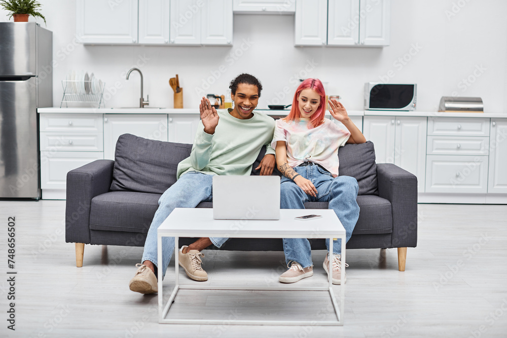 joyful beautiful multicultural couple sitting on sofa and waving at laptop camera during video call