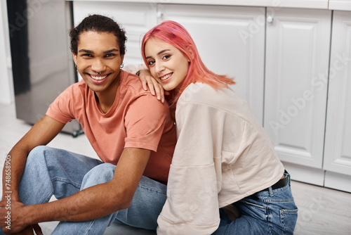 joyous appealing diverse couple in homewear sitting on floor and smiling at camera while at home
