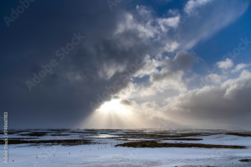 Dramatic winter landscape in Iceland with white and black clouds over snowy and icy hilly landscape with bright sun between clouds and sunrays on shining on white snow landscape