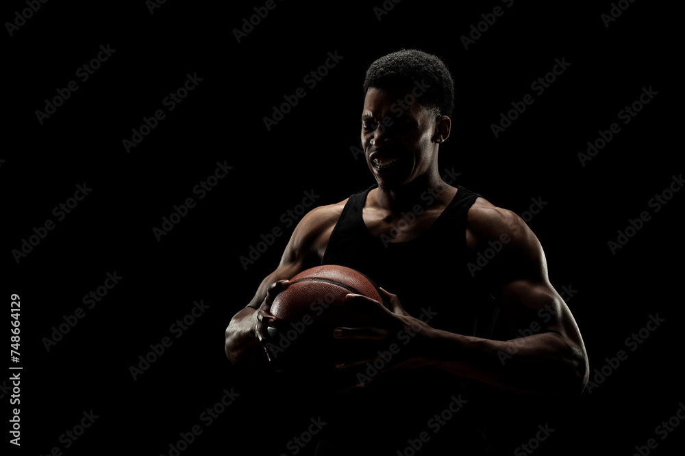 Basketball player holding a ball against black background. Screaming african american man silhouette.