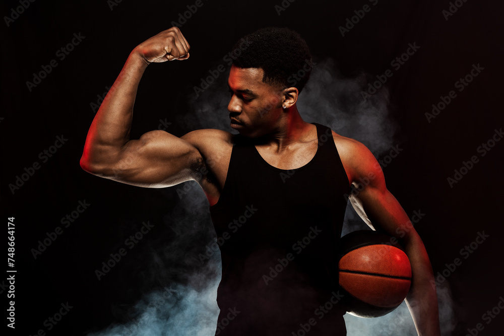 Basketball player side lit with red color holding a ball against hazy smoke fog background. Serious african american man showing muscles Basketball player side lit with red color holding a ball against hazy smoke fog background. Serious african american man showing muscles