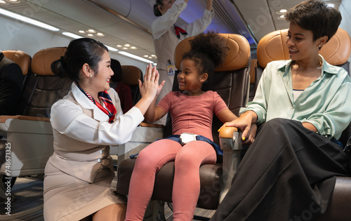 Asian air hostess providing service to passengers in airplane cabin. Smiling female flight attendant giving high five to happiness African little girl in curly hair after helping her to fasten seat be photo