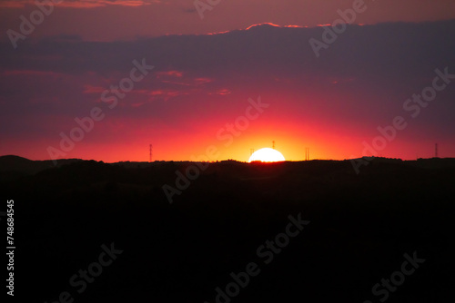Sol poniéndose en el horizonte con un color rojizo intenso. Horizonte compuesto por colinas sobre las que se encuentran pilones de la red  eléctrica. photo