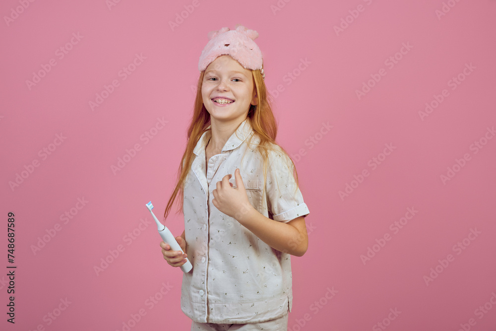 Smiling redhead teen girl holding toothbrush and wearing sleeping mask on pink background.