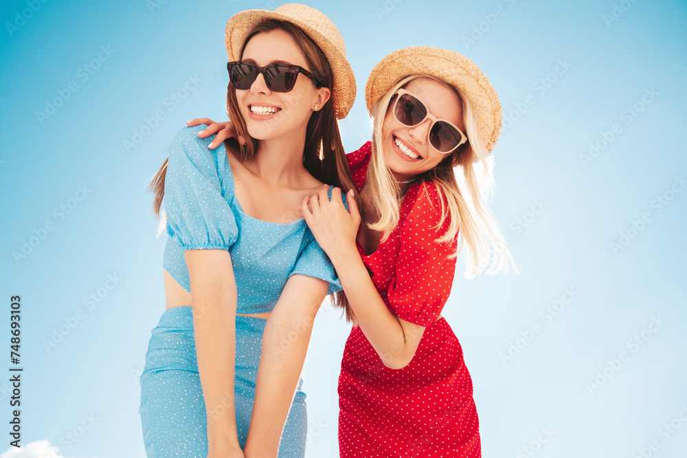 Two young beautiful smiling hipster woman in trendy summer same clothes. Carefree women in street in hats. Positive models at sunset. Cheerful and happy. Bottom view. They look at camera from above Two young beautiful smiling hipster woman in trendy summer same clothes. Carefree women in street in hats. Positive models at sunset. Cheerful and happy. Bottom view. They look at camera from above