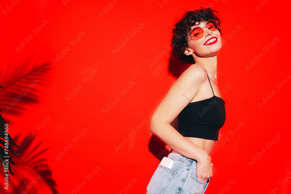 Young beautiful smiling female in trendy summer black tank top. Carefree woman posing near red wall in studio with curly hairstyle. Positive model having fun. Cheerful and happy. In sunglasses Young beautiful smiling female in trendy summer black tank top. Carefree woman posing near red wall in studio with curly hairstyle. Positive model having fun. Cheerful and happy. In sunglasses