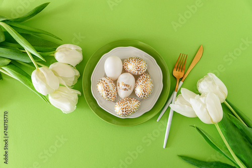 Plate decorated golden Easter eggs and white tulips on green background. Easter celebration concept. Flat lay. Top view
