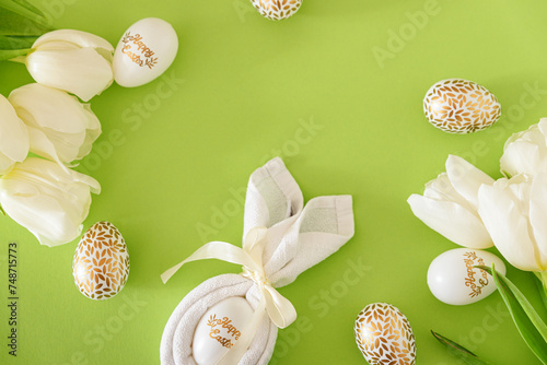 Flat lay with  bunny ears napkin, white tulips and Easter eggs on green background. Easter celebration concept. Top view