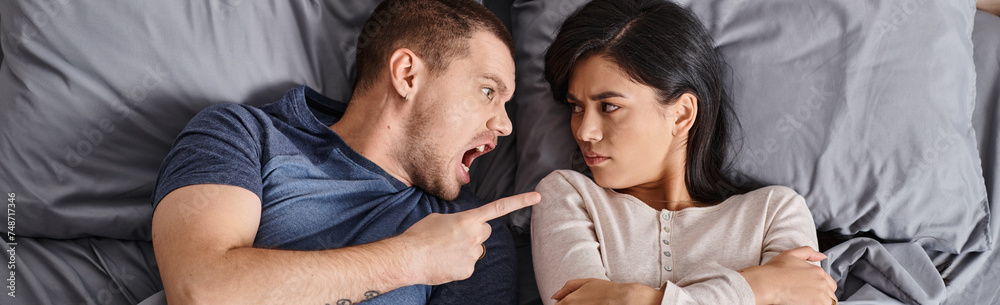 Obraz premium angry multicultural couple lying down on bed shouting and quarreling at each other, top view, banner