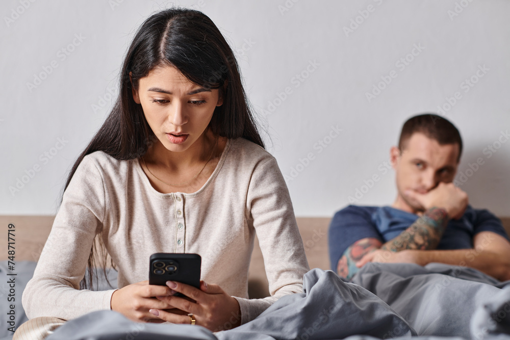 shocked asian woman looking at smartphone near husband lying on bed at home, family conflict shocked asian woman looking at smartphone near husband lying on bed at home, family conflict