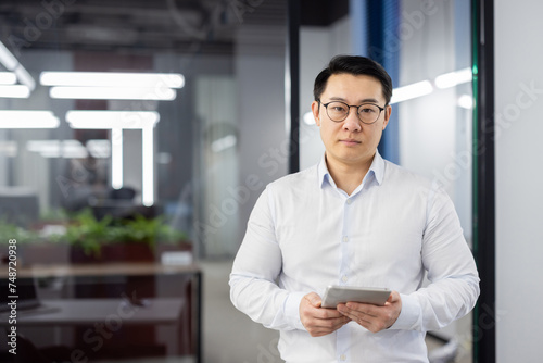 Portrait of a serious and confident Asian man standing in the office in a white shirt and glasses, holding a tablet and looking at the camera.