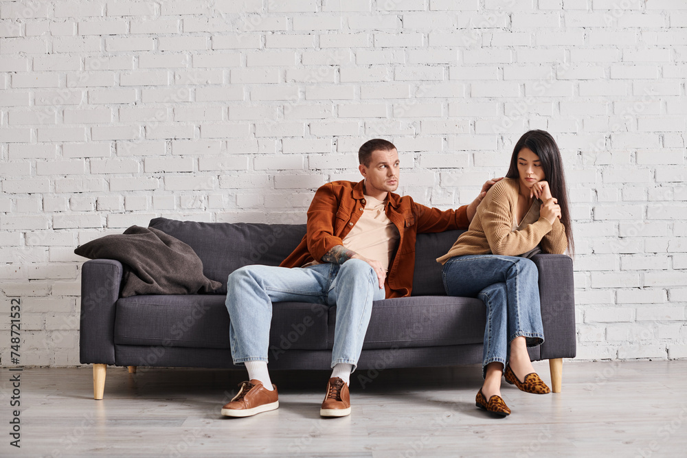 young man calming upset asian wife sitting on couch in living room, relationship difficulties young man calming upset asian wife sitting on couch in living room, relationship difficulties