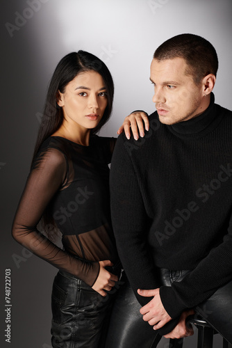 seductive asian woman near trendy man in black attire looking at camera while sitting on grey