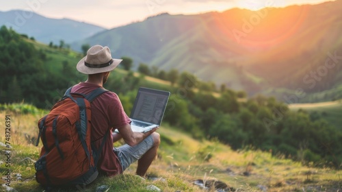 Young man freelancer traveler wearing hat anywhere working online using laptop and enjoying mountains view photo