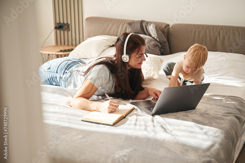 Woman in headphones with laptop, writing in notepad. Mother with her little son is at home in domestic room