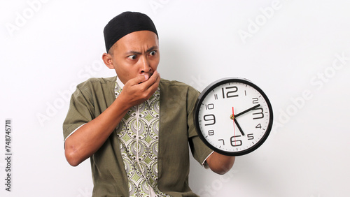 A shocked Indonesian Muslim man in koko and peci holds a clock, realizing he is late for his sahur meal during Ramadan. Isolated on a white background photo