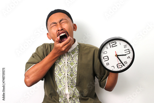 Sleepy Indonesian Muslim man in koko and peci stifles a yawn while checking a clock. He has just woken up to prepare for the suhoor meal before the Ramadan fast begins. Isolated on a white background photo