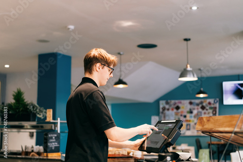 Young waiter serving customer at cash point in cafe. Man working with POS terminal. Cashier, barista checking for payment receipt. Hospitality, server and preparing a slip at the till in coffee shop