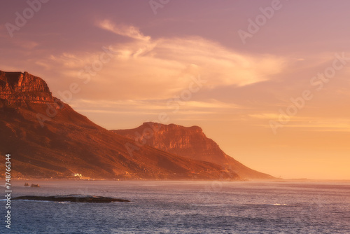 Ocean, sunset and clouds on mountains by blue sky and outdoor travel for vacation in nature. Landscape, sea and sunlight on false bay with calm and seal island in cape town for tourist destination photo