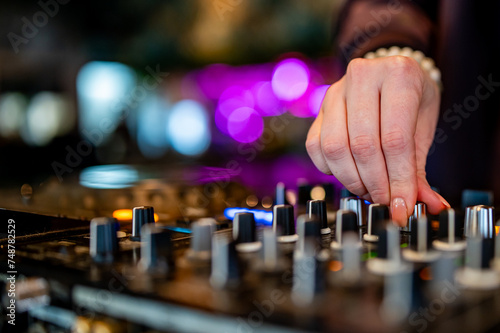 close-up of hands skillfully adjusting knobs and sliders on a professional DJ mixer, illuminated by colorful lights. The mood conveys focus and control during a live music event