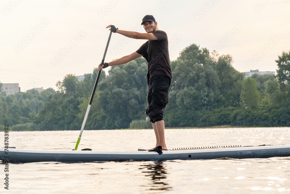 Naklejka premium Young man has fun gliding on paddle board over urban lake. Sport enthusiast spends vacation engaged in sports activities