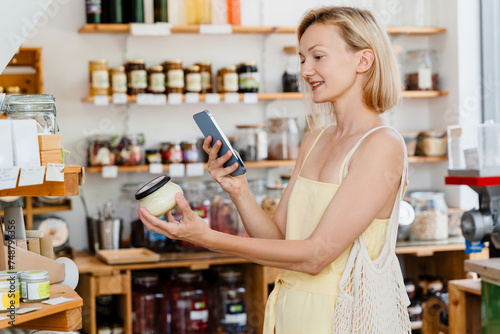 Thoughtful woman using smartphone buying products at store. Сonscious shopping and retail of organic food and eco goods at local small shop.