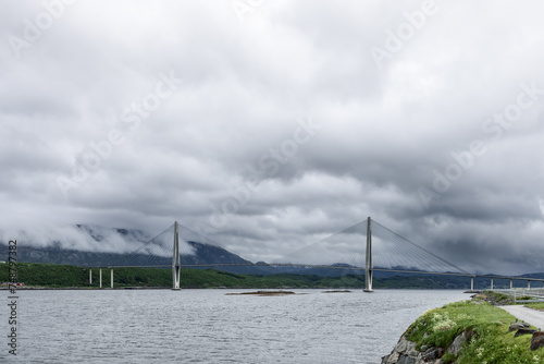 Lines of a suspension bridge on the Kystriksveien route in Norway frame a view shrouded in clouds, showcasing a blend of engineering and mist-laden nature photo
