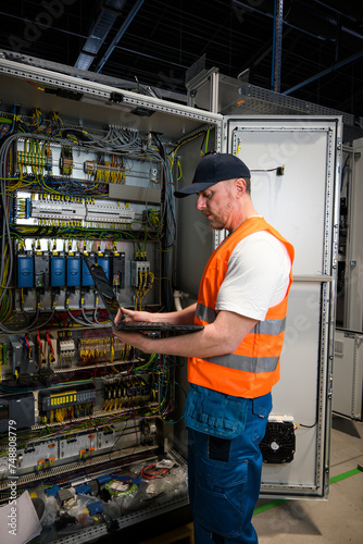 an electrician in a cap and orange vest installs a large electrical substation using a computer