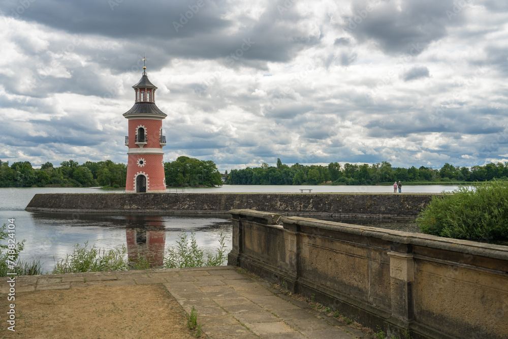 An elderly couple, a man and a woman, enjoy a beautiful view of a pink lighthouse against the backdrop of dark, stormy clouds. Lighthouse near Moritzburg, Germany