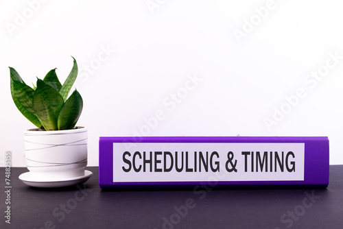 Folder with the text label Scheduling and Timing lies on a dark table with a flower and a light background. photo