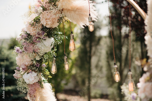 The wedding arch is decorated with flowers of white and lilac shades, pampas grass and a garland with retro lamps, romantic atmosphere, close-up. Photo zone for a couple on Valentine's Day or a weddin