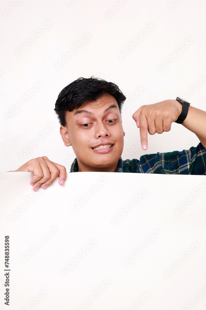 Portrait of young asian man standing behind an empty signboard and pointing on it Portrait of young asian man standing behind an empty signboard and pointing on it