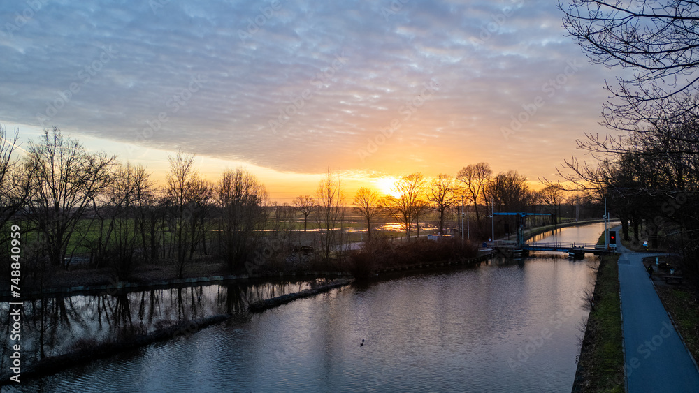 This serene image captures a sunset by a calm canal, with the sun dipping low in the sky and casting a vibrant orange hue along the horizon. The silhouette of leafless trees reflects on the water's This serene image captures a sunset by a calm canal, with the sun dipping low in the sky and casting a vibrant orange hue along the horizon. The silhouette of leafless trees reflects on the water's